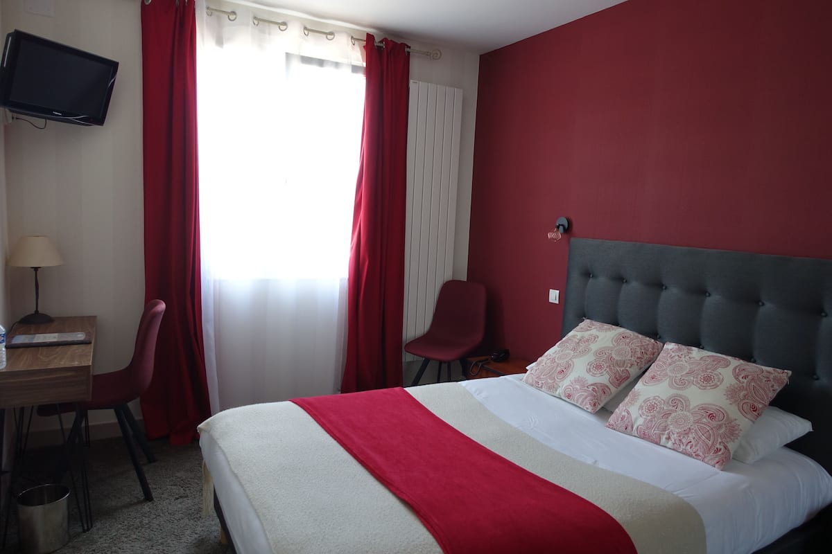 Une chambre d’hôtel avec un lit, un bureau, une chaise, une télévision et une fenêtre avec des rideaux.