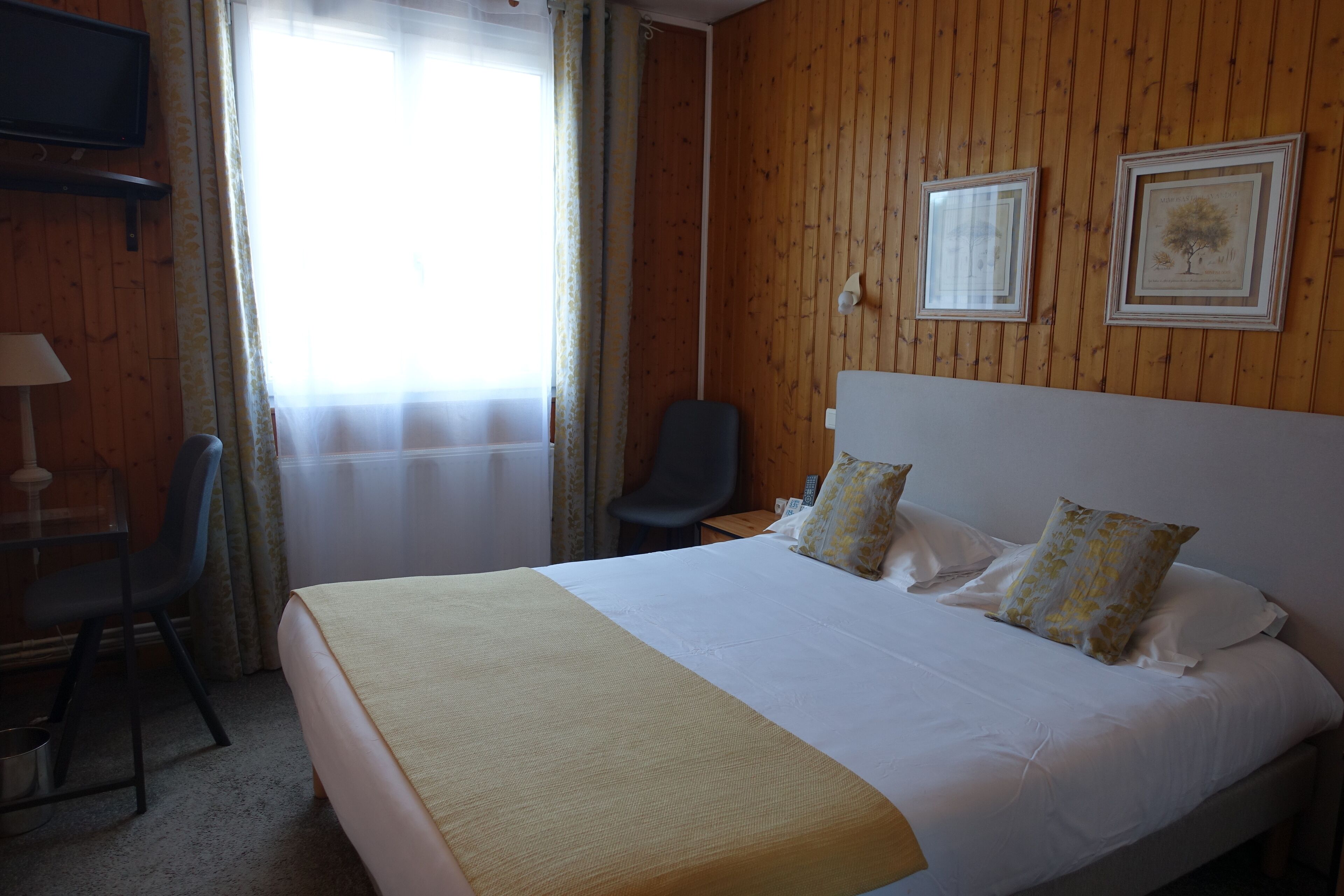 A hotel room with a large bed, a desk with a chair, a television mounted on the wall, and two framed pictures on the wall.