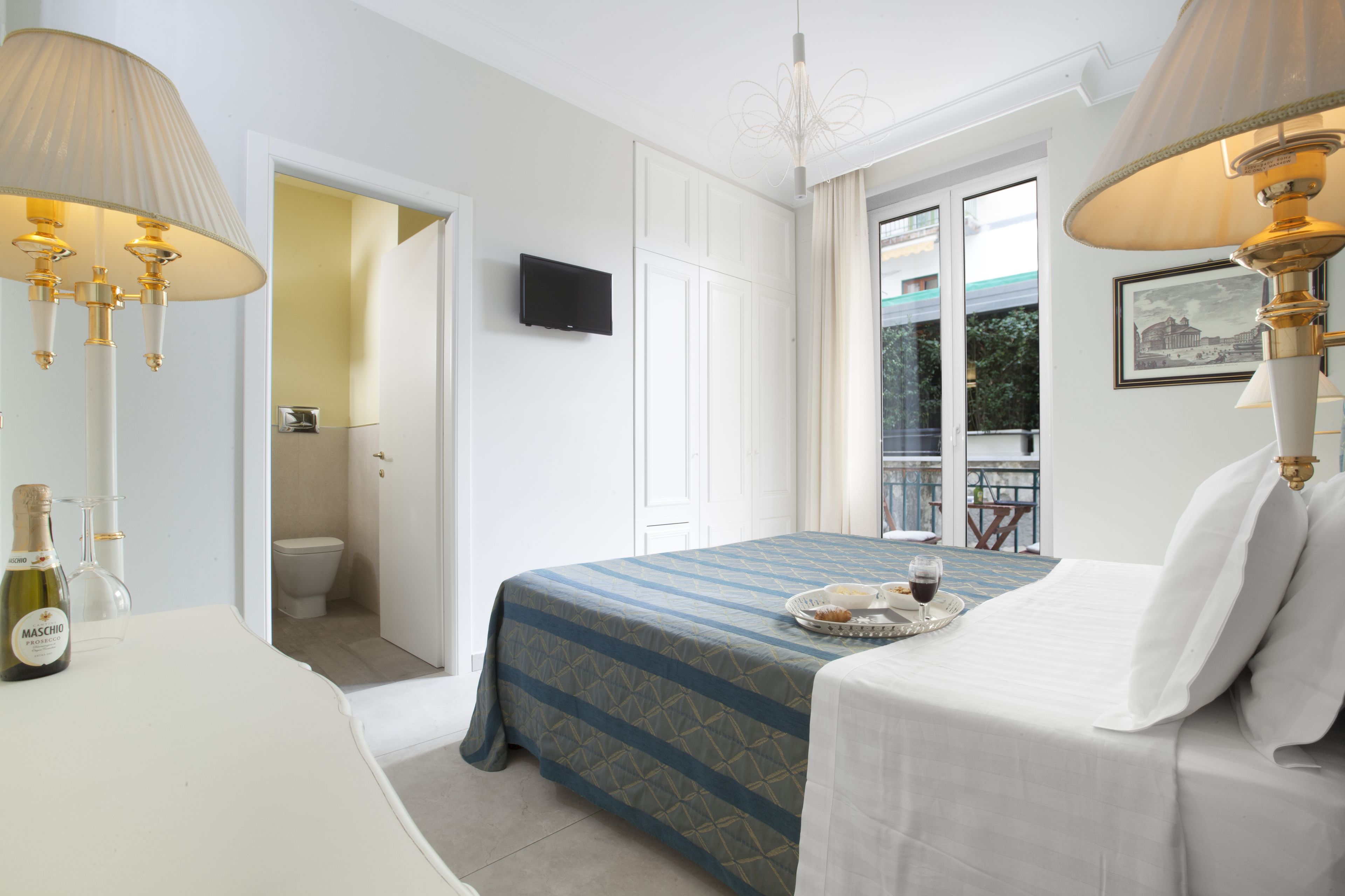 A hotel room with a bed, bedside lamps, a television, a minibar, and a balcony view.
