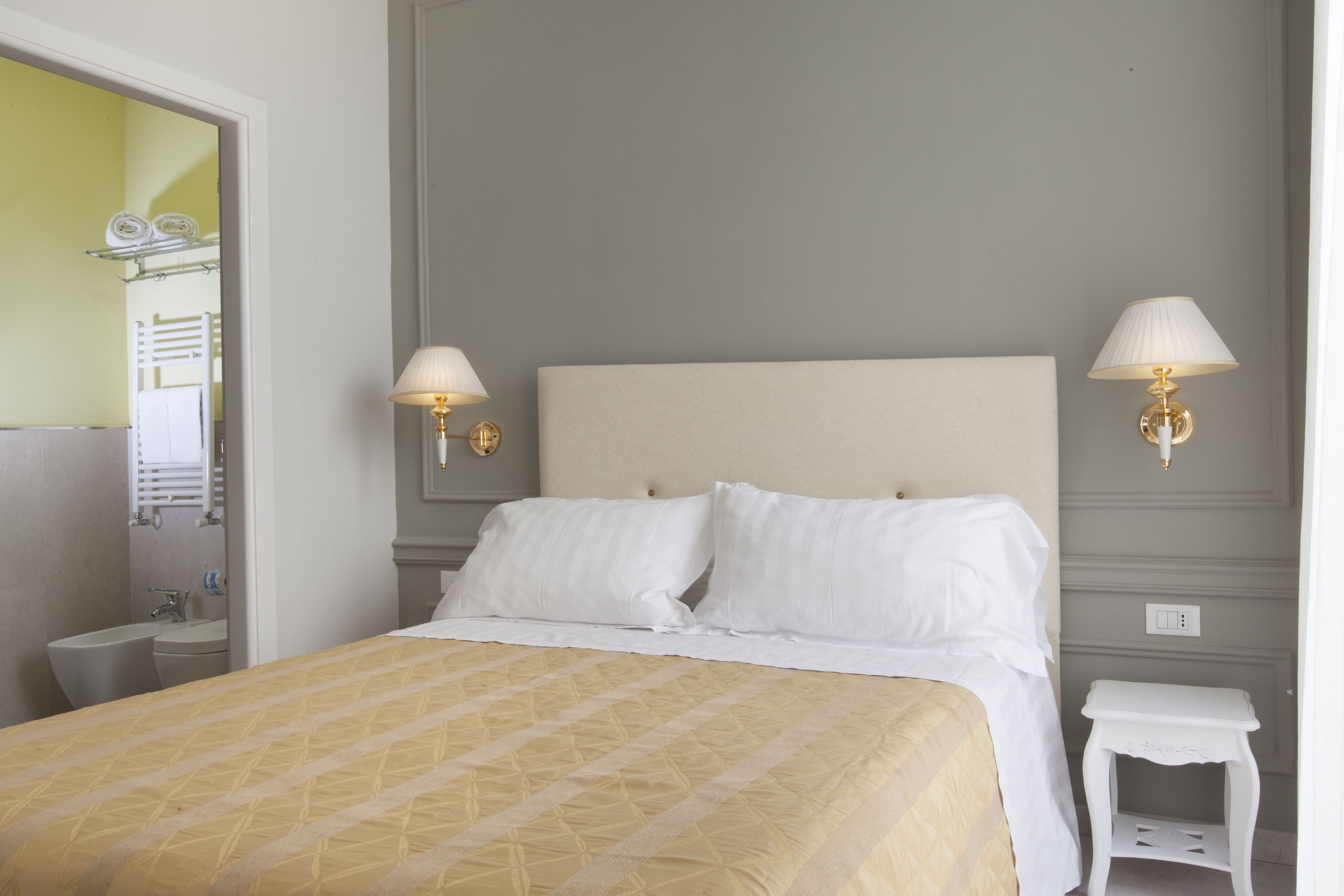 Een netjes opgemaakte hotelkamer met een bed, bedlampjes, een klein wit nachtkastje en een spiegel die de badkamer weerspiegelt.
