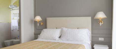 A neatly arranged hotel room with a bed, bedside lamps, a small white nightstand, and a mirror reflecting the bathroom.
