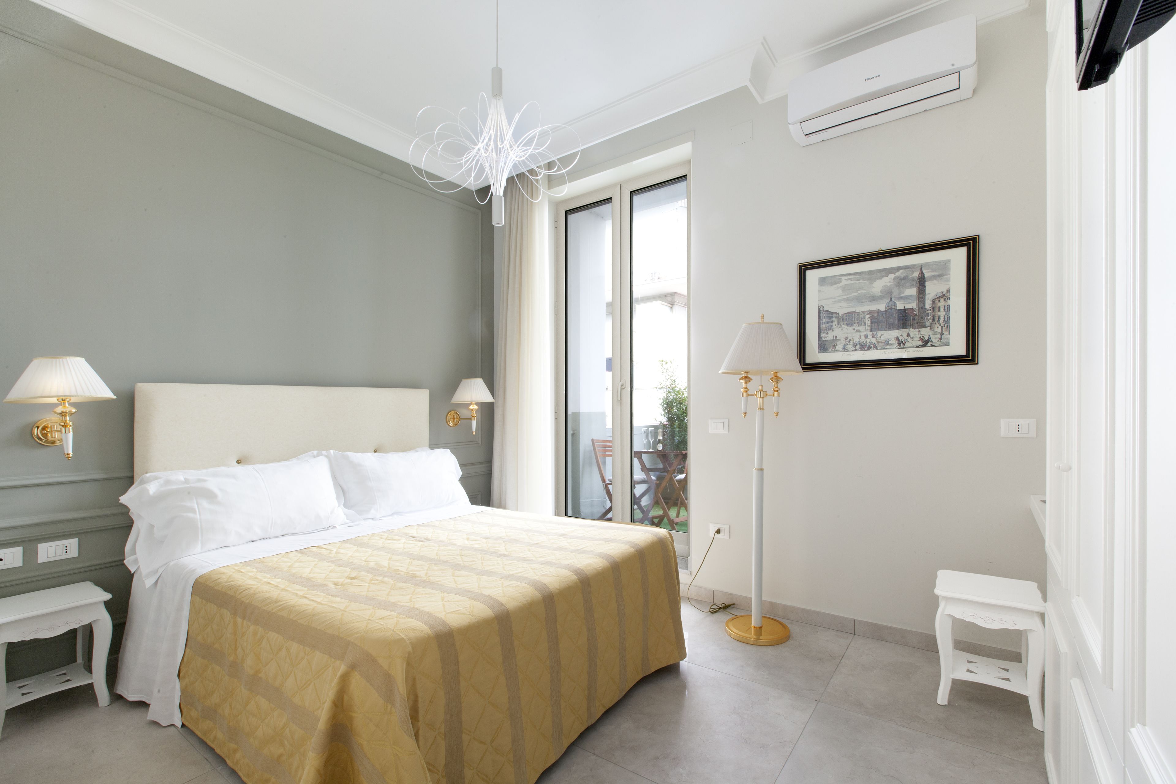 Ein Hotelzimmer mit Bett, Nachttischen, einer Chandelüster, einer Klimaanlage, einem Bild an der Wand und Blick durch das Fenster.