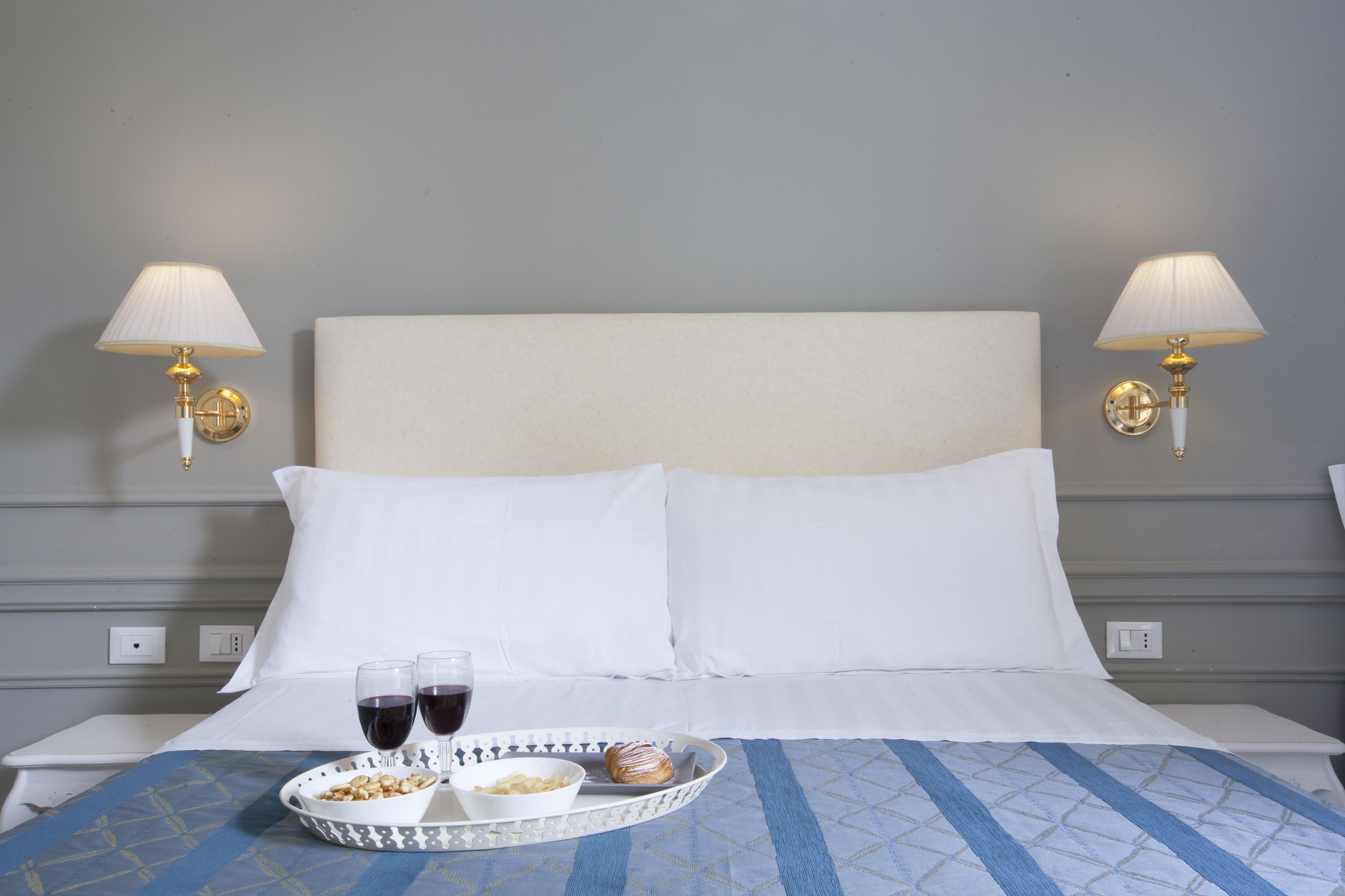 A hotel room with a bed, two bedside lamps, a tray with snacks, and two glasses of red wine.