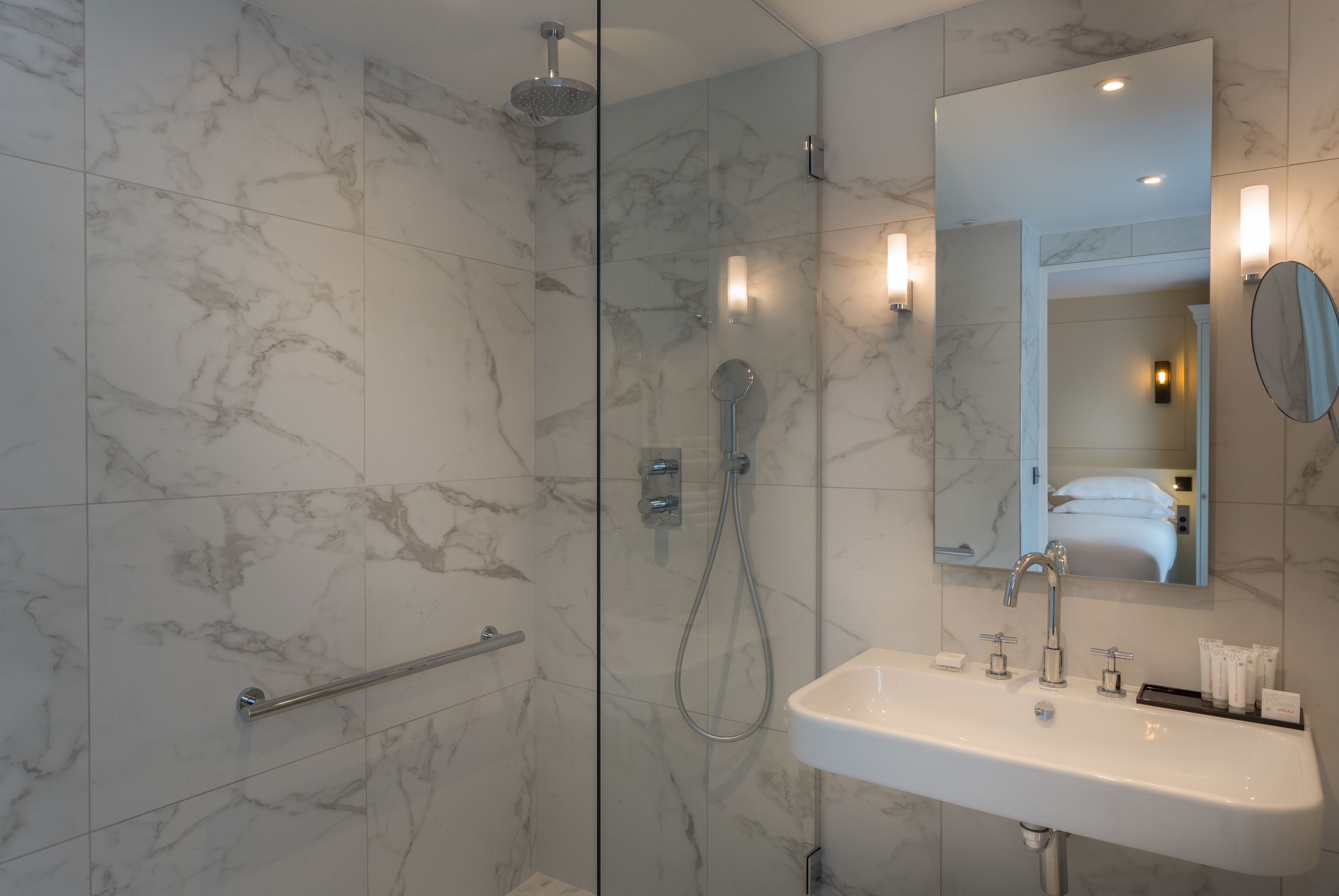 A modern bathroom with a glass-enclosed shower, a white sink with toiletries, and a large mirror.