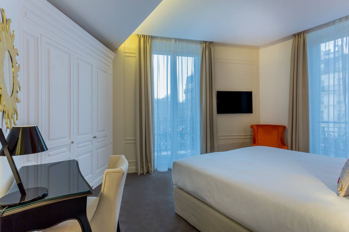 Une chambre d’hôtel avec un lit, un bureau doté d’une lampe, une chaise, un téléviseur fixé au mur et de grandes fenêtres avec des rideaux.