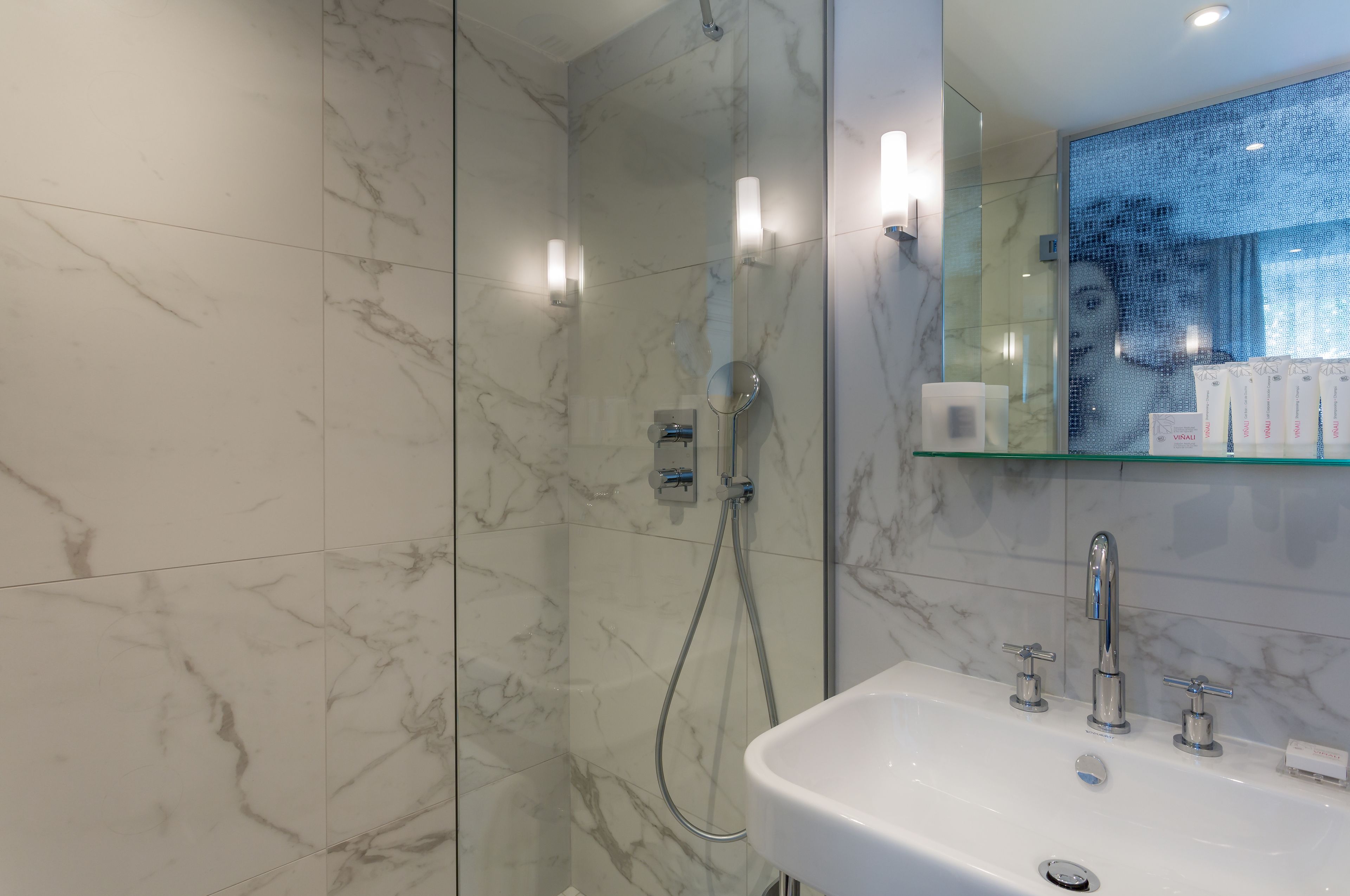 Une salle de bain moderne avec un mur de douche en marbre, une étagère en verre où sont rangés les articles de toilette et un lavabo blanc avec des robinets chromés.