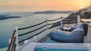A poolside area with a view of the sea and a sunset, featuring a hot tub, seating, and a table with drinks. - Windmill Luxury Villa (Santorini)