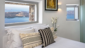 A bedroom with a large window offering a view of the sea and cliffs, a framed painting above the bed, and a small window with a view of the sea. - Windmill Luxury Villa (Santorini)