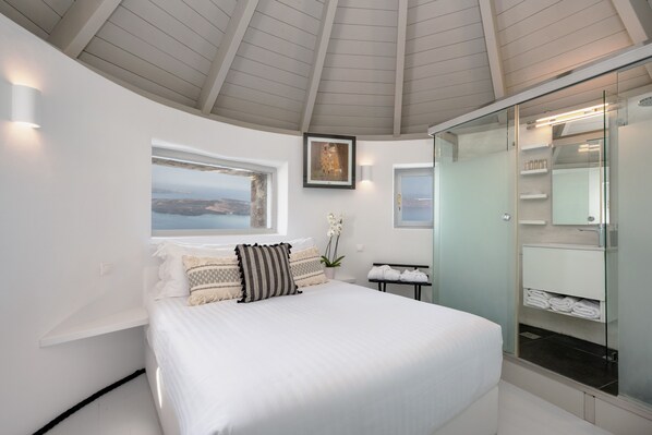 A modern bedroom with a large bed, a view of the sea, a glass shower enclosure, and a framed artwork on the wall.