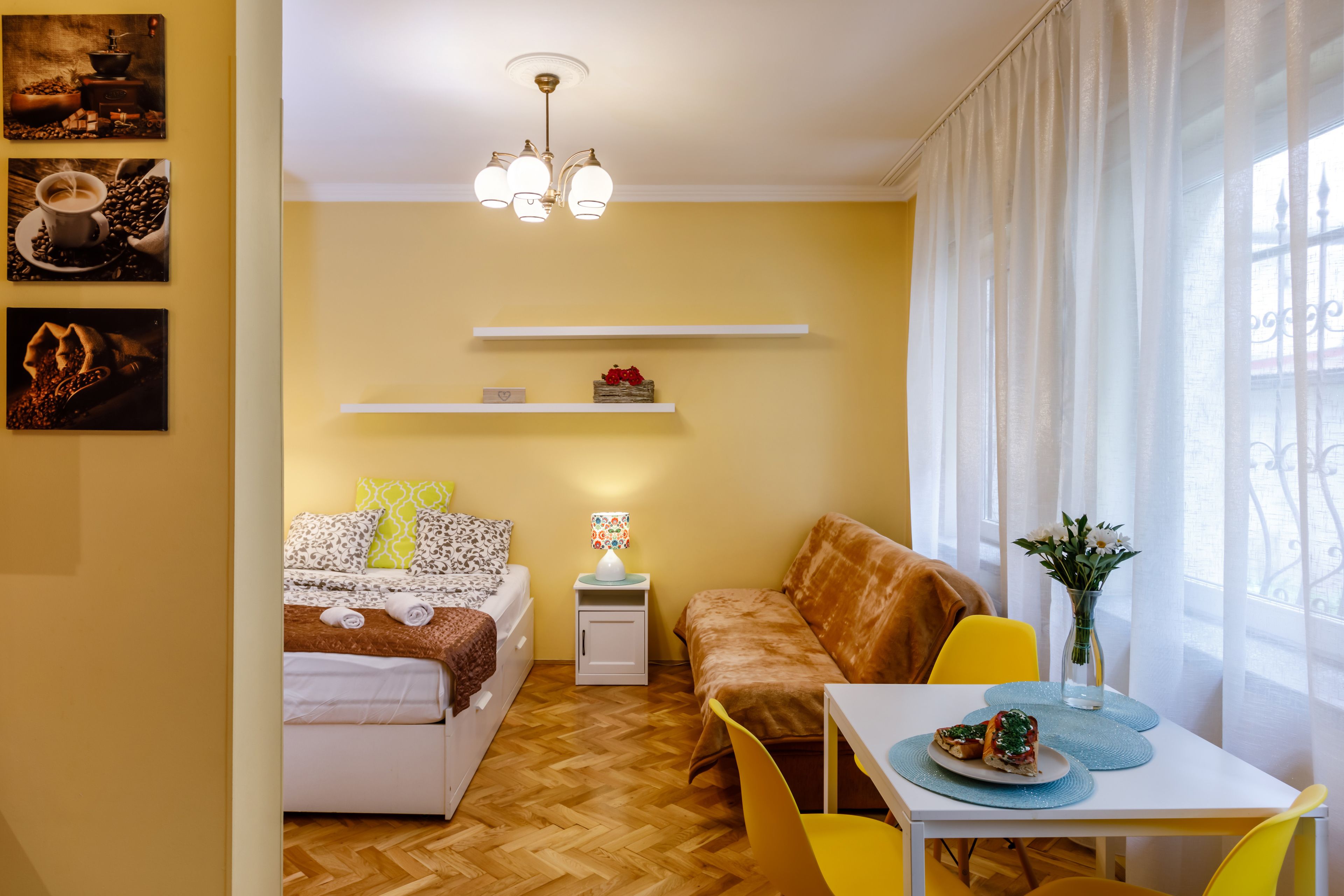A compact hotel room with a bed, a sofa, a dining table with chairs, a small side table, and a window with curtains.