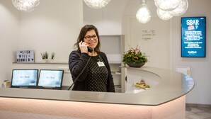 Een receptiebalie met een modern design, met een gebogen toonbank en hangende pendelverlichting. Twee computermonitoren staan op de balie, waarop 'Go.Bed' te zien is. Een bord met 'SBAR' is rechts op de muur zichtbaar.
