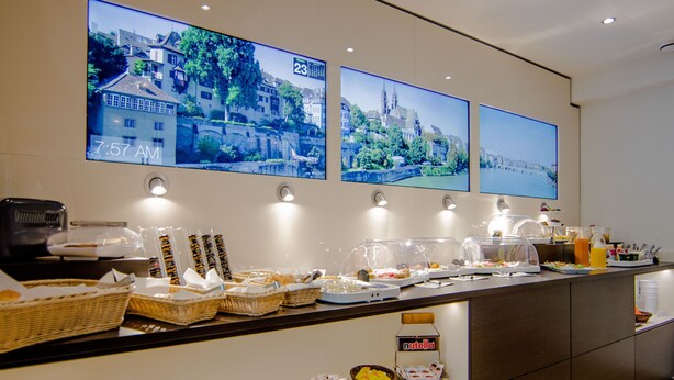 Een ontbijtbuffet met diverse etenswaren en manden, een klok waarop 7:57 uur staat, en drie ingelijste foto’s aan de muur.