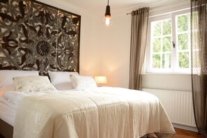 A neatly made bed with a decorative headboard, a bedside lamp, and a window with curtains. - VillaAdel Designroom (Alsfeld)