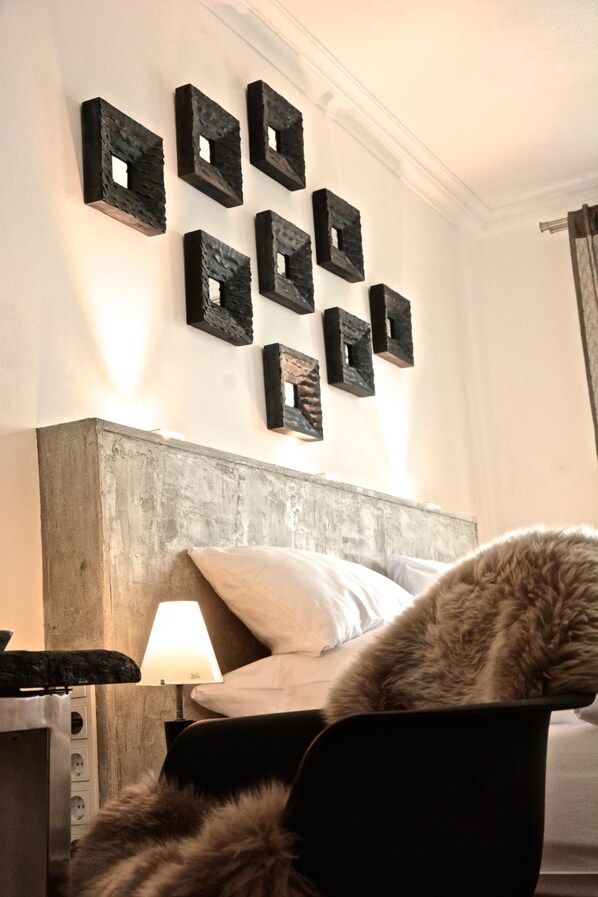 Un dormitorio con cabecera de madera, cama con ropa blanca, lámpara sobre una mesita de noche y un gato sobre una silla.