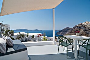 Een terras met een witte tafel en stoelen, planten in potten en uitzicht op zee en bergen.