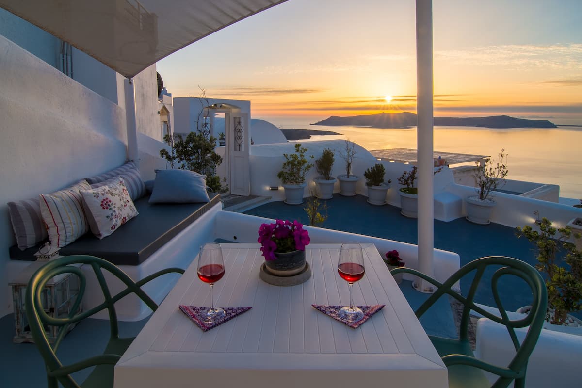 A terrace with a white table, two chairs, and a sofa with cushions. There are potted plants and a vase with flowers on the table.