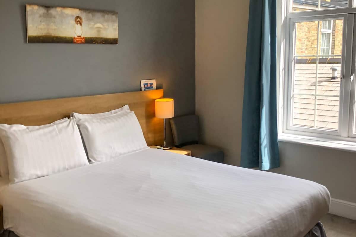 Cama bem arrumada com roupa de cama branca, cabeceira de madeira, luminária de cabeceira e janela com cortinas.