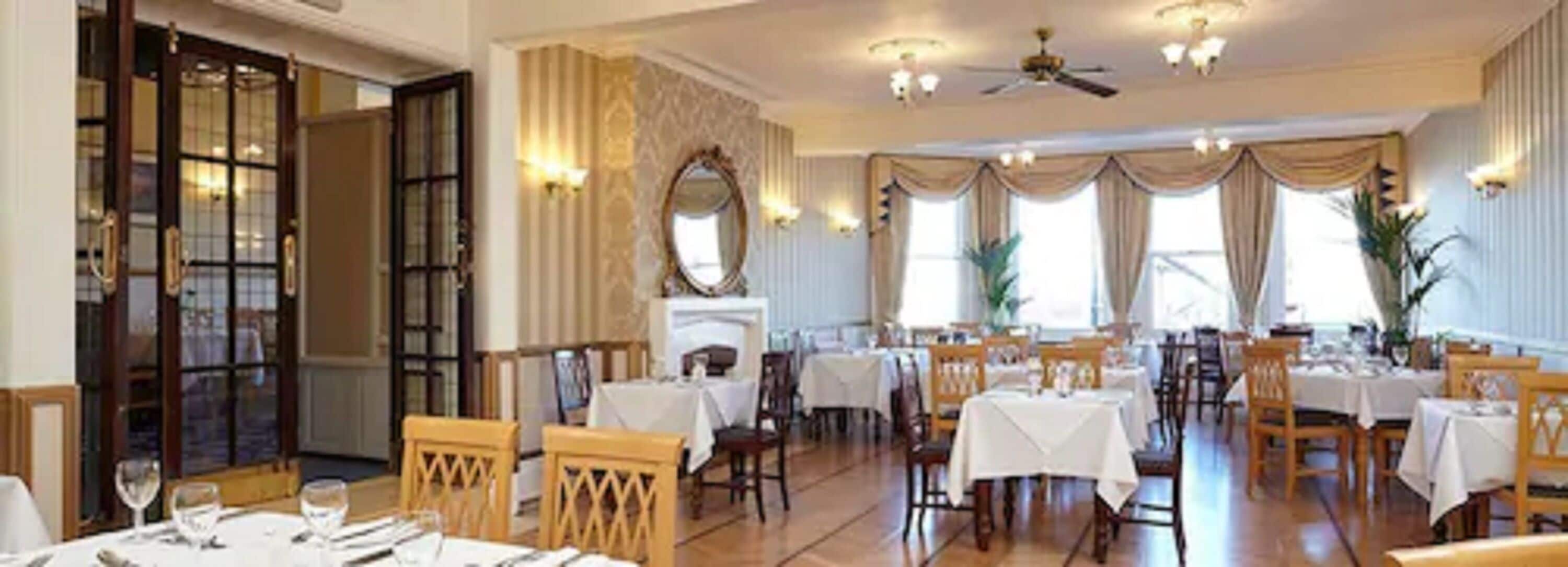 A dining area with multiple tables and chairs, large windows with curtains, and a ceiling fan.