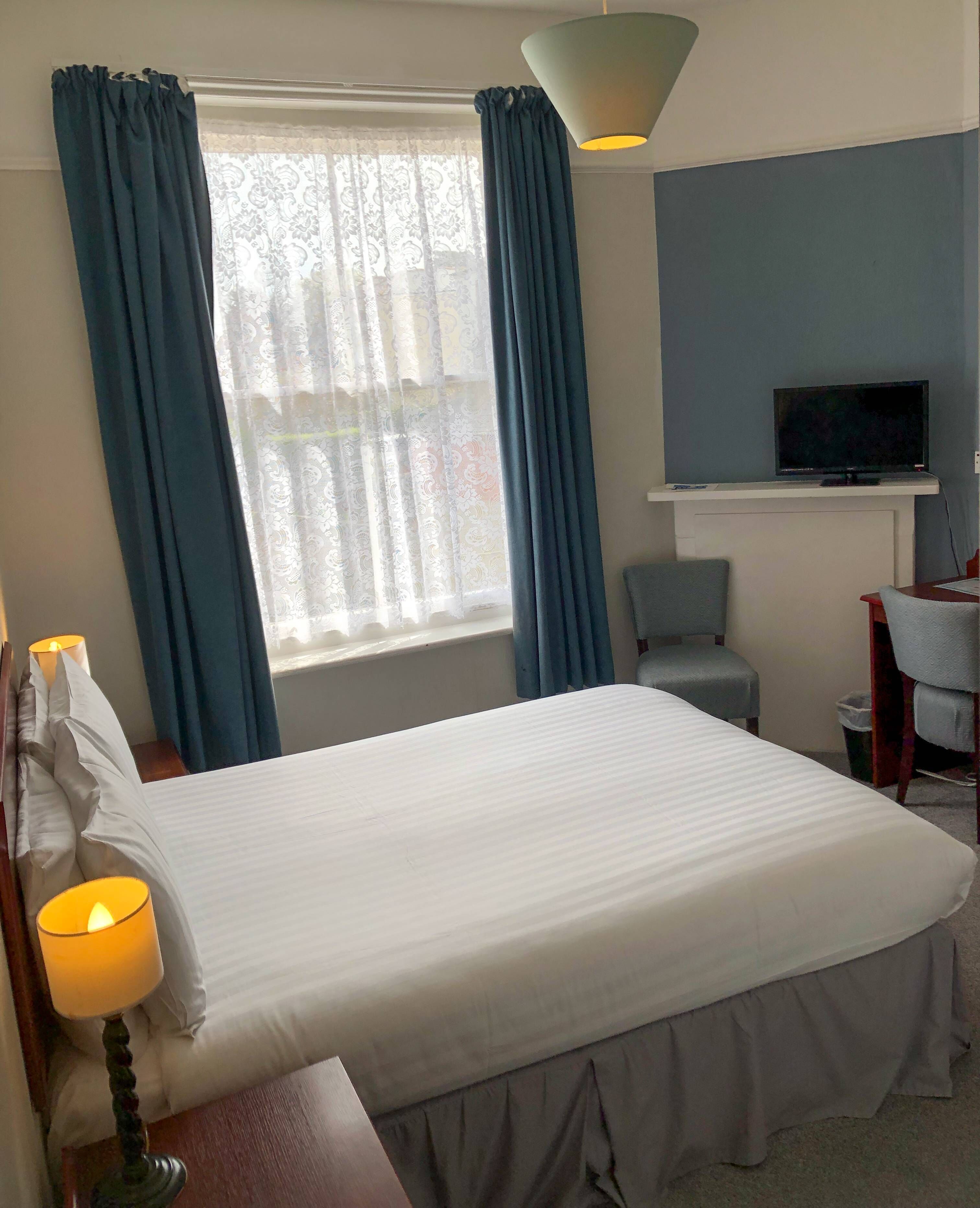 A hotel room with a bed, a desk with a chair, a television, and a window with curtains.