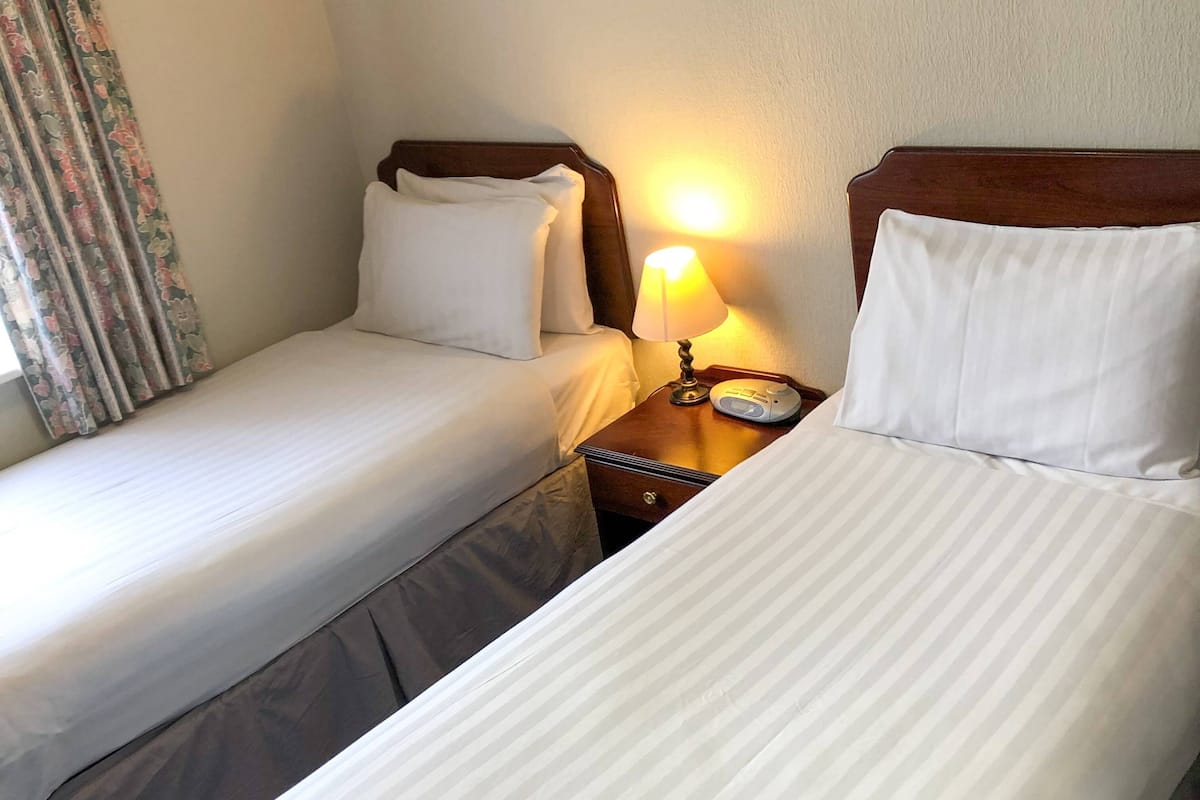 Duas camas de solteiro com cabeceiras de madeira, mesa de cabeceira com abajur e cortina florida.