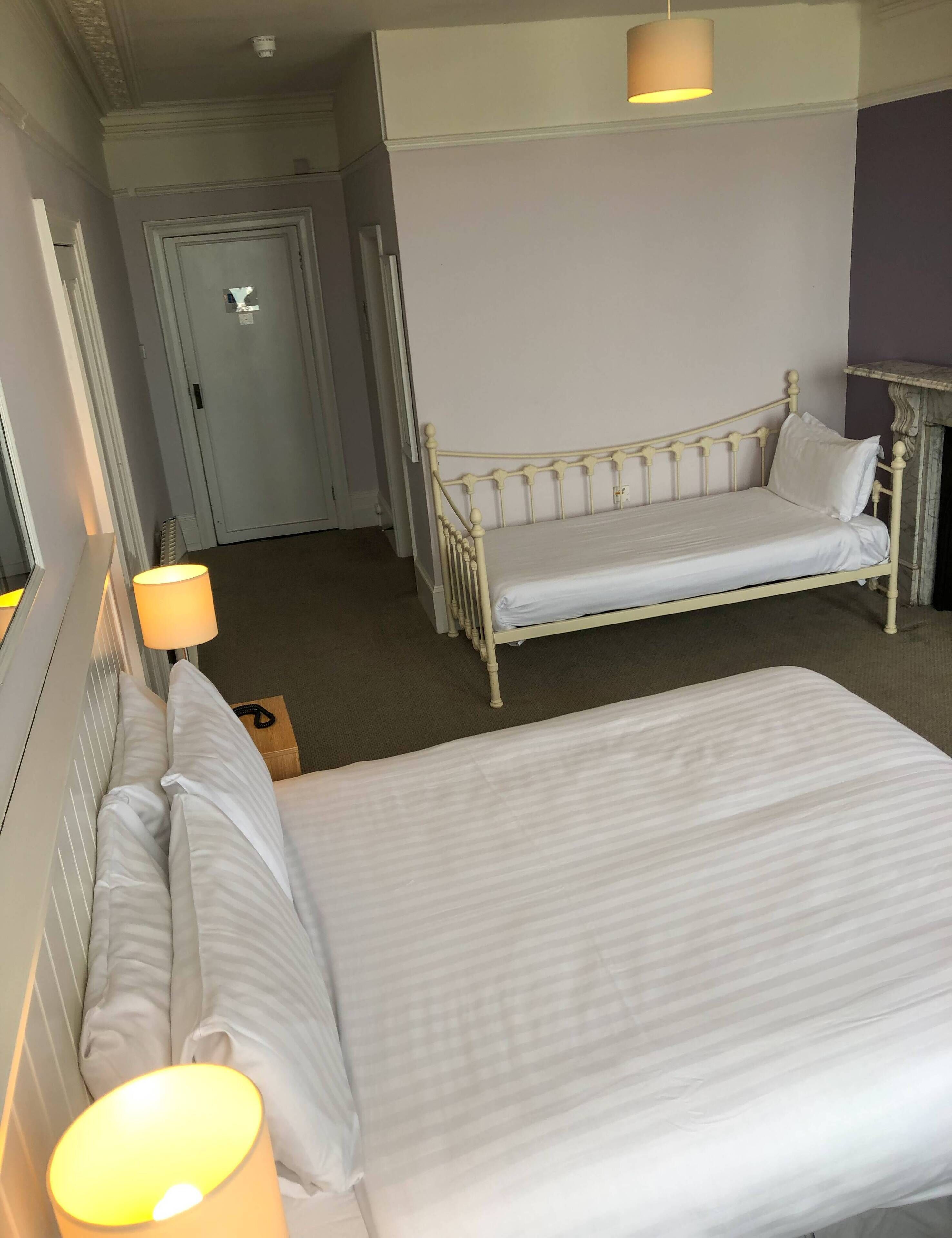 A hotel room with two beds, a door, a lamp, and a fireplace.