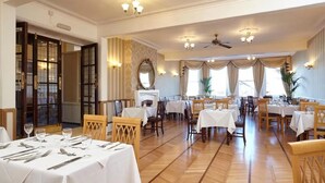Un amplio comedor con varias mesas y sillas, chimenea y ventanas grandes con cortinas.