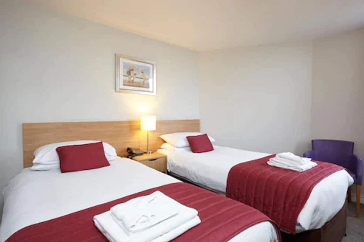 Quarto de hotel com duas camas, uma mesa de cabeceira, uma luminária, uma cadeira e um quadro na parede.
