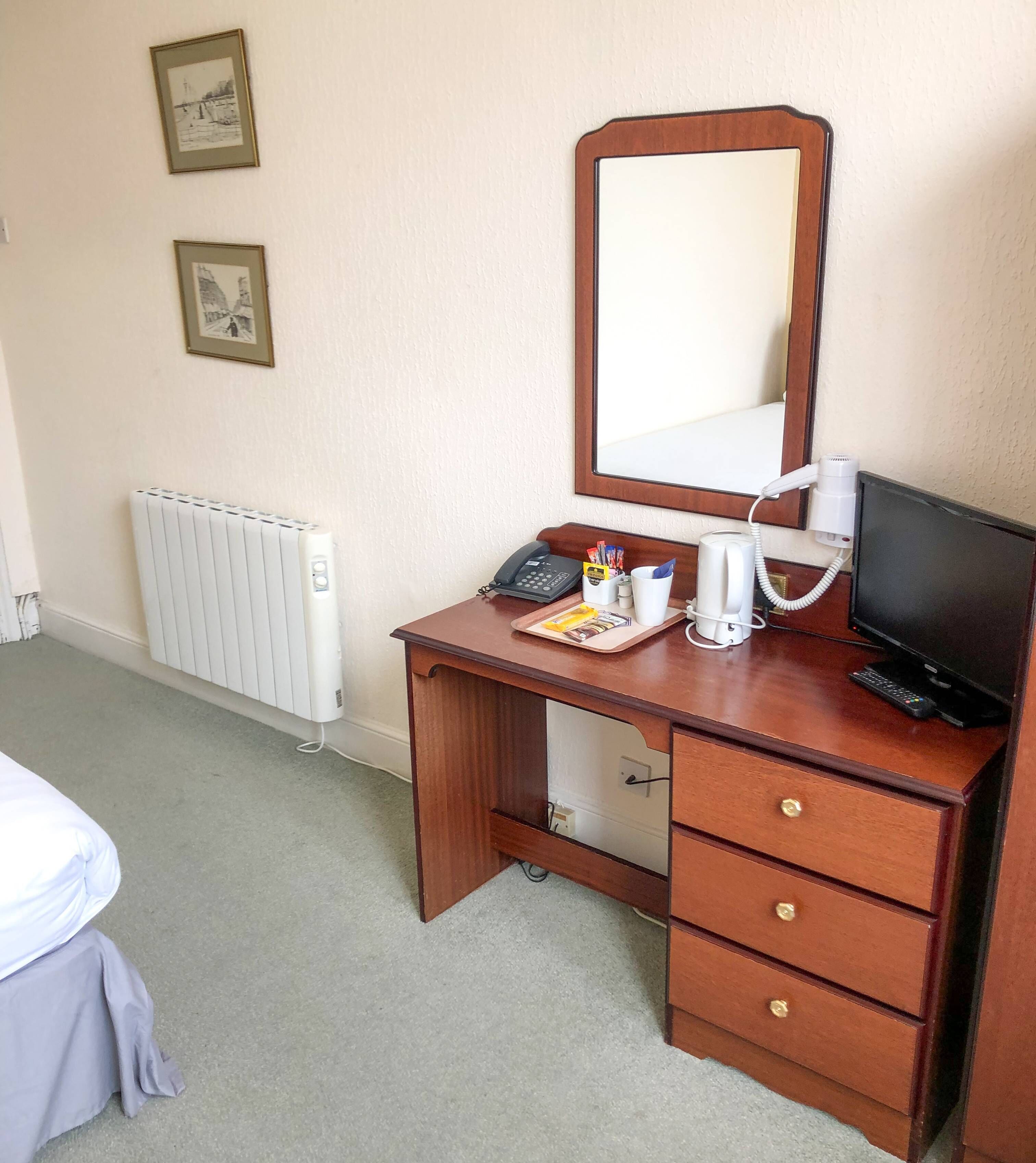 A wooden desk with a mirror, a telephone, a coffee maker, and a television.