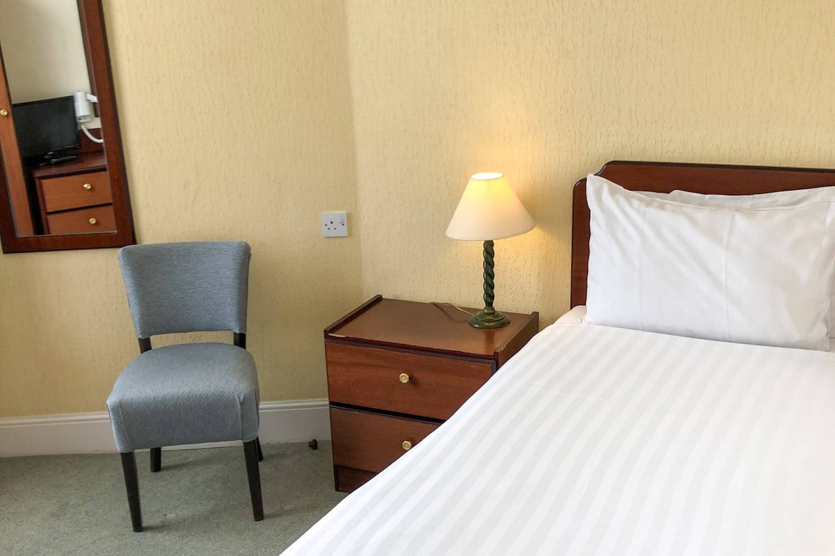 Quarto de hotel com cama, uma cadeira, mesa de cabeceira com abajur e cômoda.