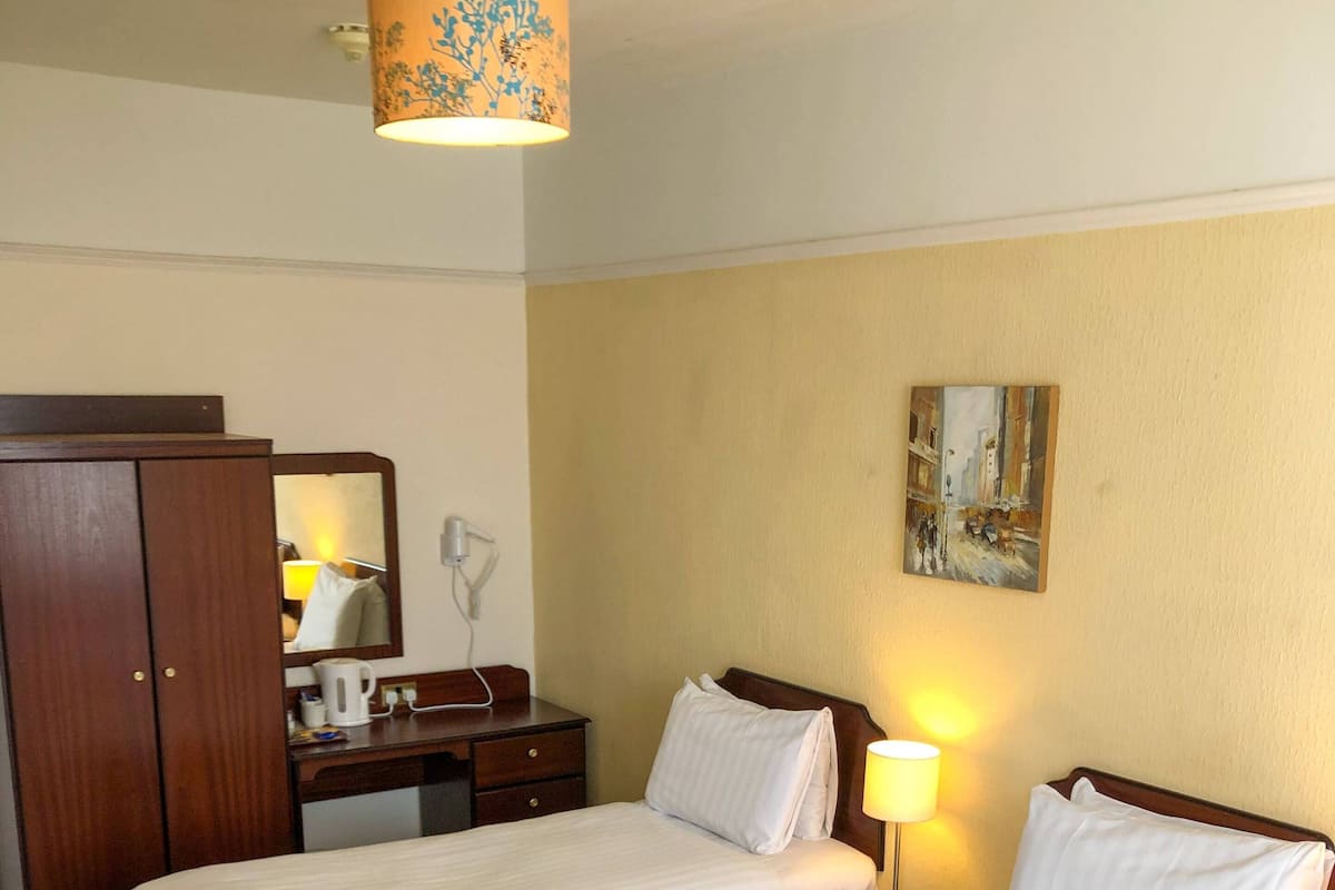 Quarto de hotel com duas camas de solteiro, um guarda-roupa de madeira, uma escrivaninha com abajur e um quadro na parede.