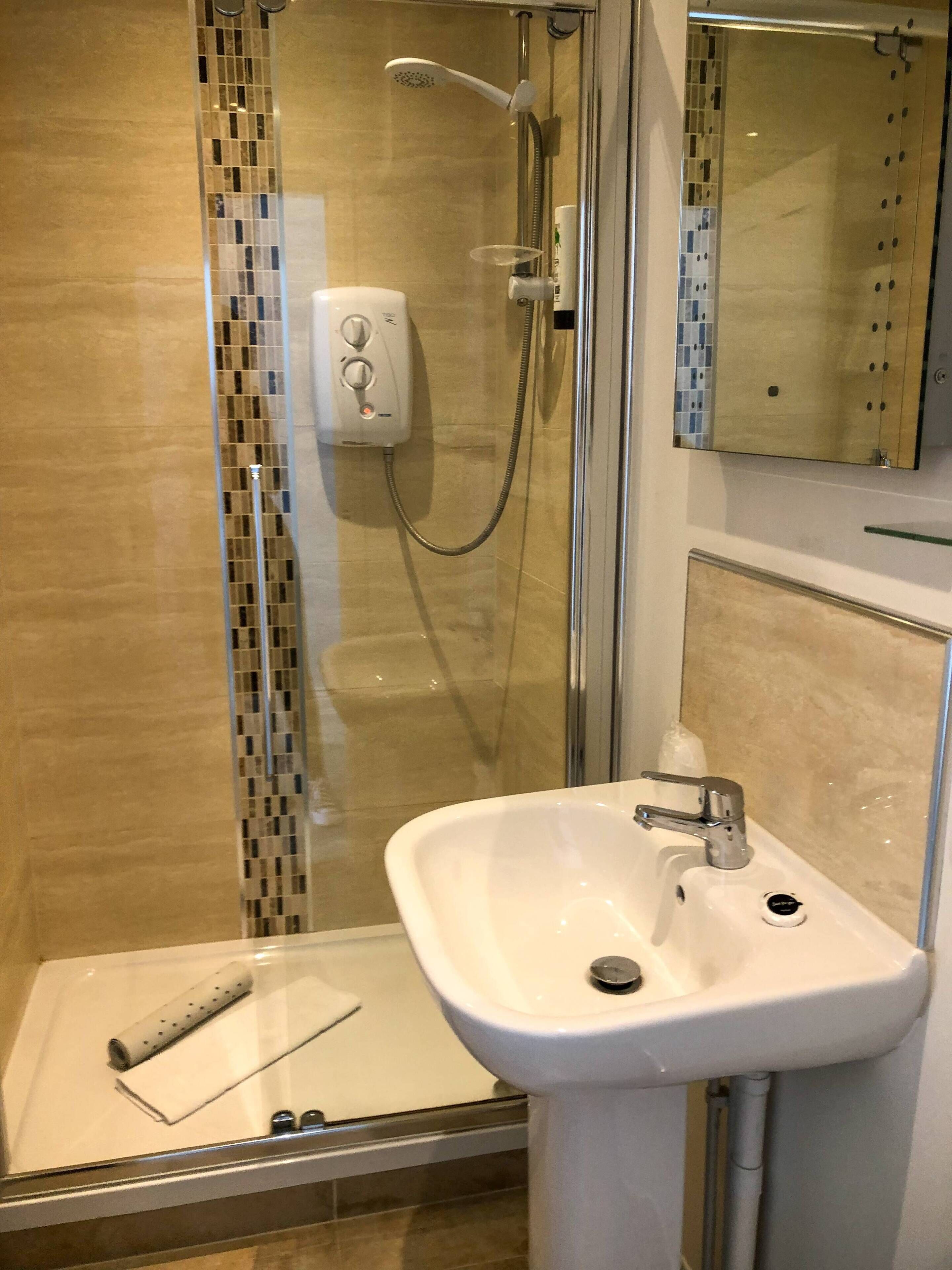 Une salle de bain moderne avec une douche à parois de verre, un lavabo blanc doté d’un seul robinet et une petite serviette posée sur le sol.