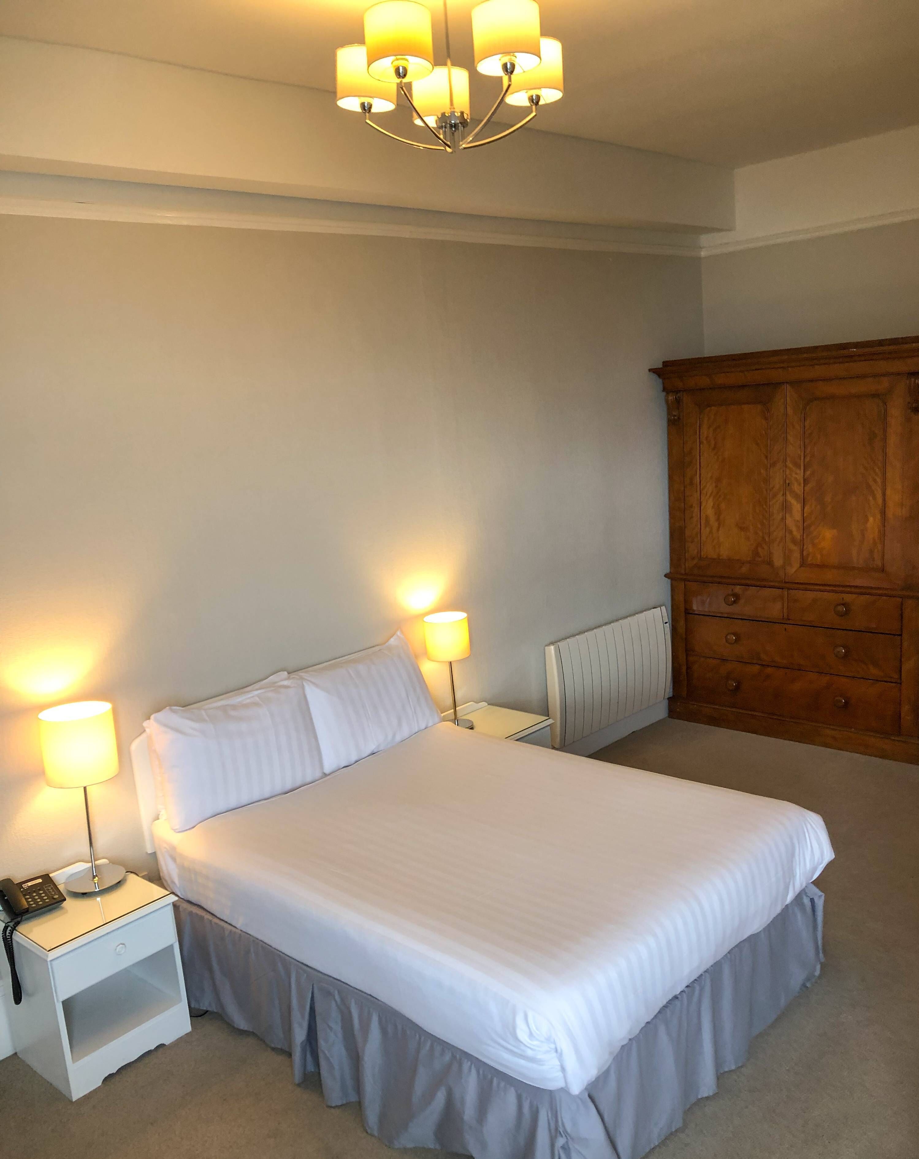 A neatly made bed in a hotel room with a wooden wardrobe, two bedside lamps, and a ceiling light fixture.