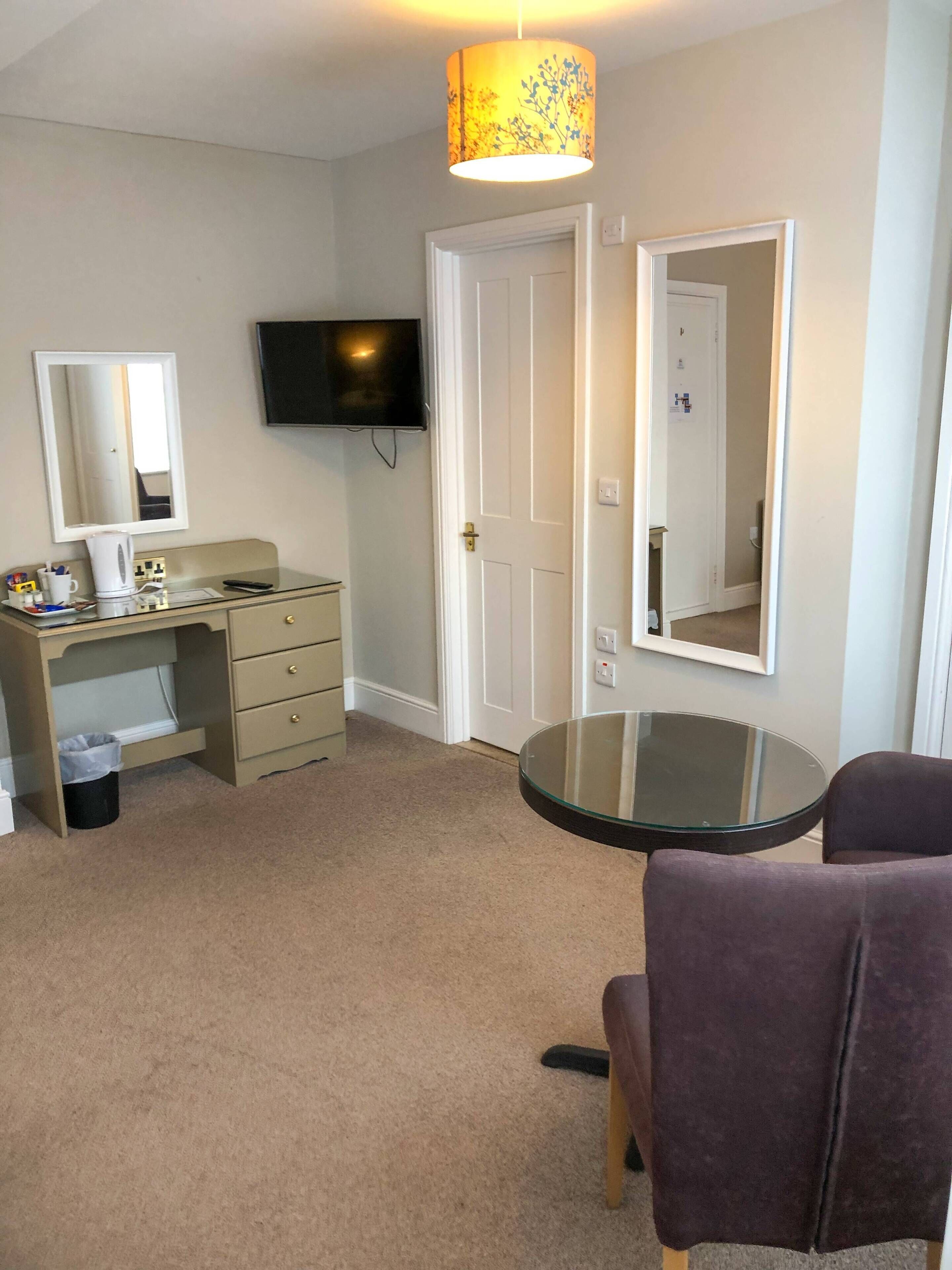 A hotel room with a beige carpet, a desk with a mirror, a television mounted on the wall, and a round glass table with two purple chairs.