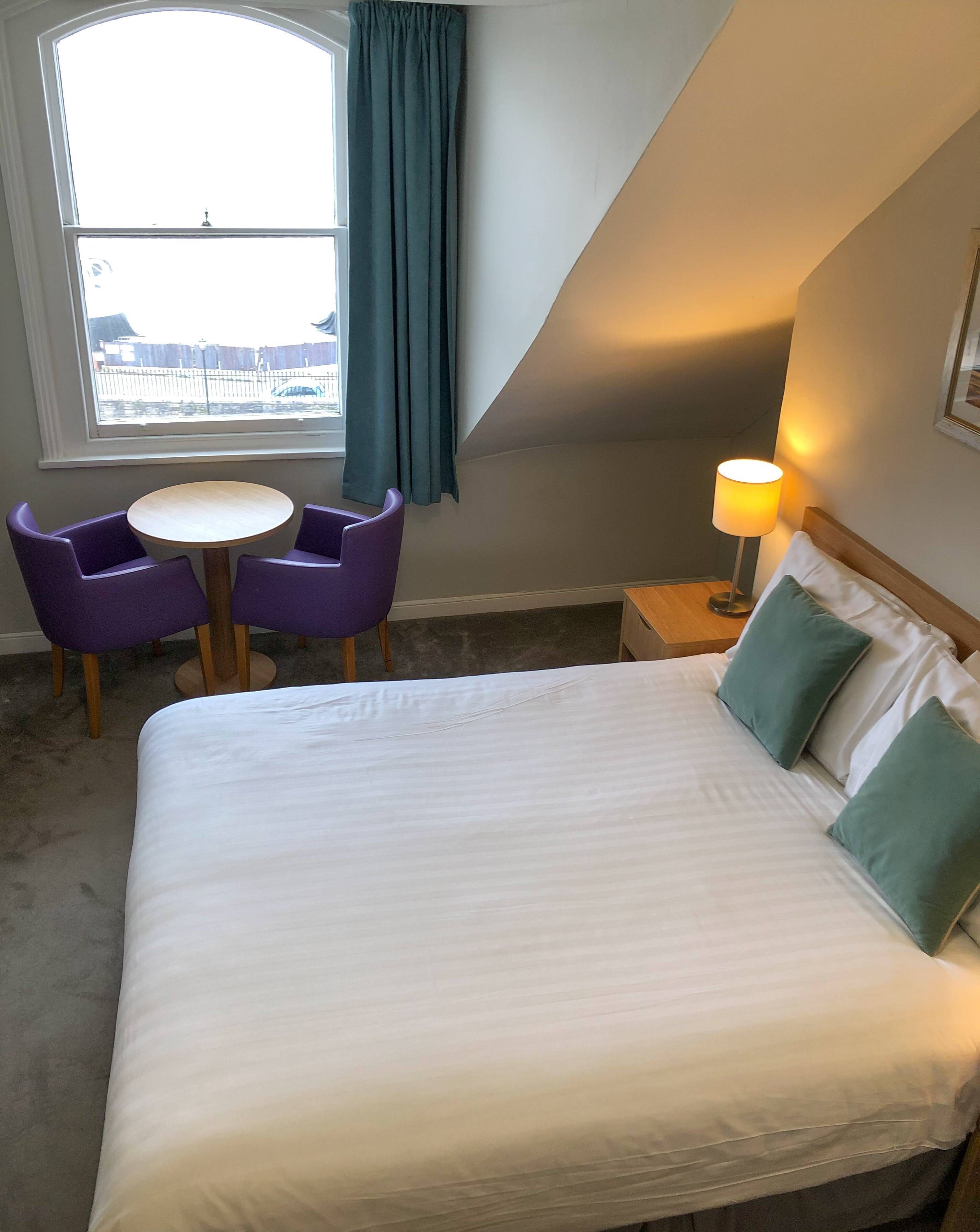 Ein Hotelzimmer mit einem Bett, zwei violetten Sessel, einem kleinen runden Tisch, einer Lampe und Blick auf ein Gewässer durch ein Fenster.
