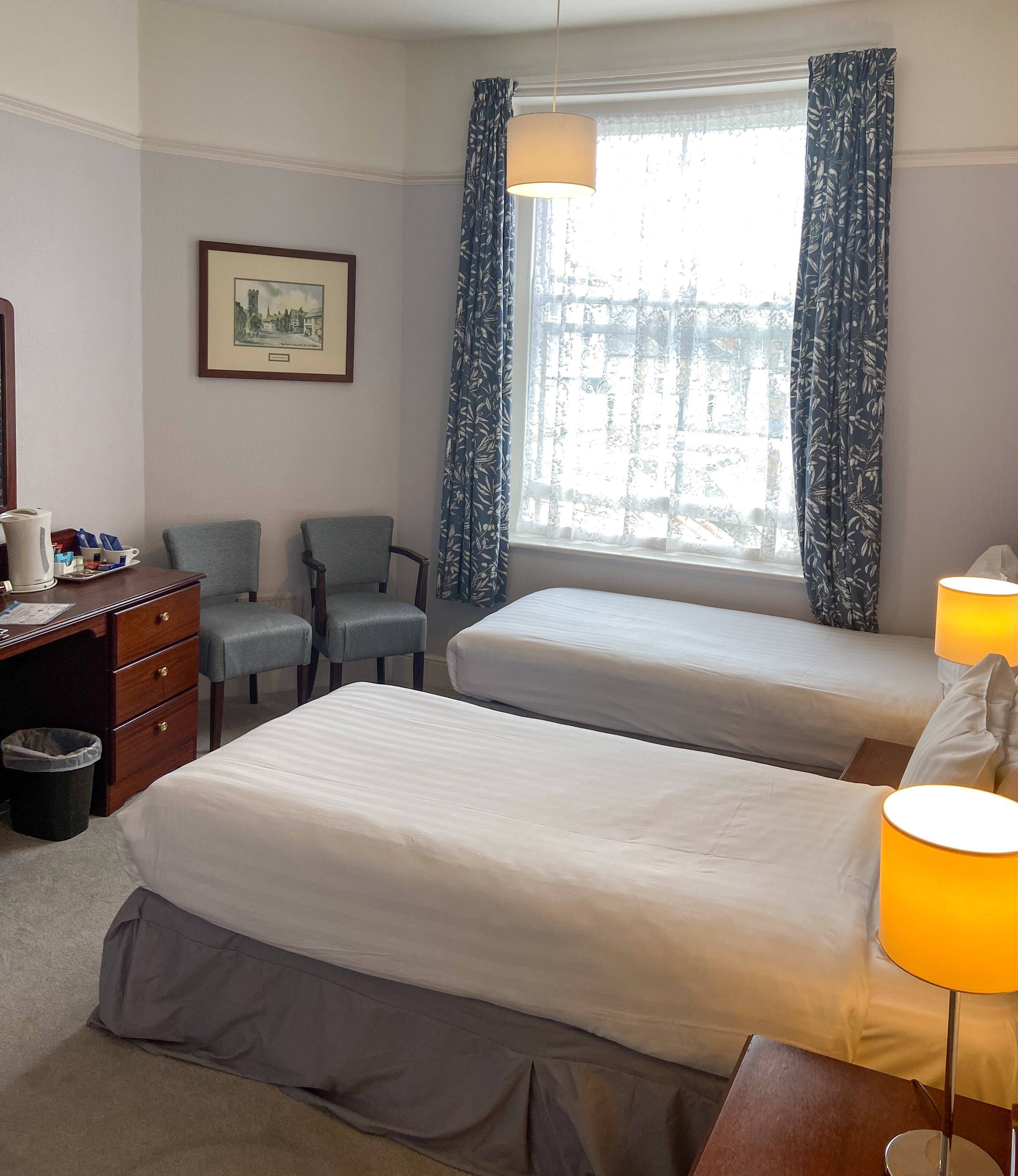 A hotel room with two beds, a desk with a lamp, a chair, a framed picture on the wall, and a window with curtains.
