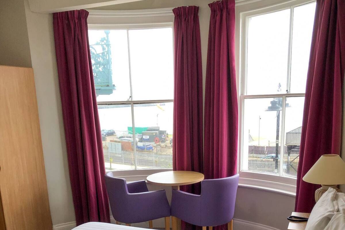 Quarto de hotel com uma cama, duas cadeiras roxas, uma mesinha redonda e uma janela com cortinas.