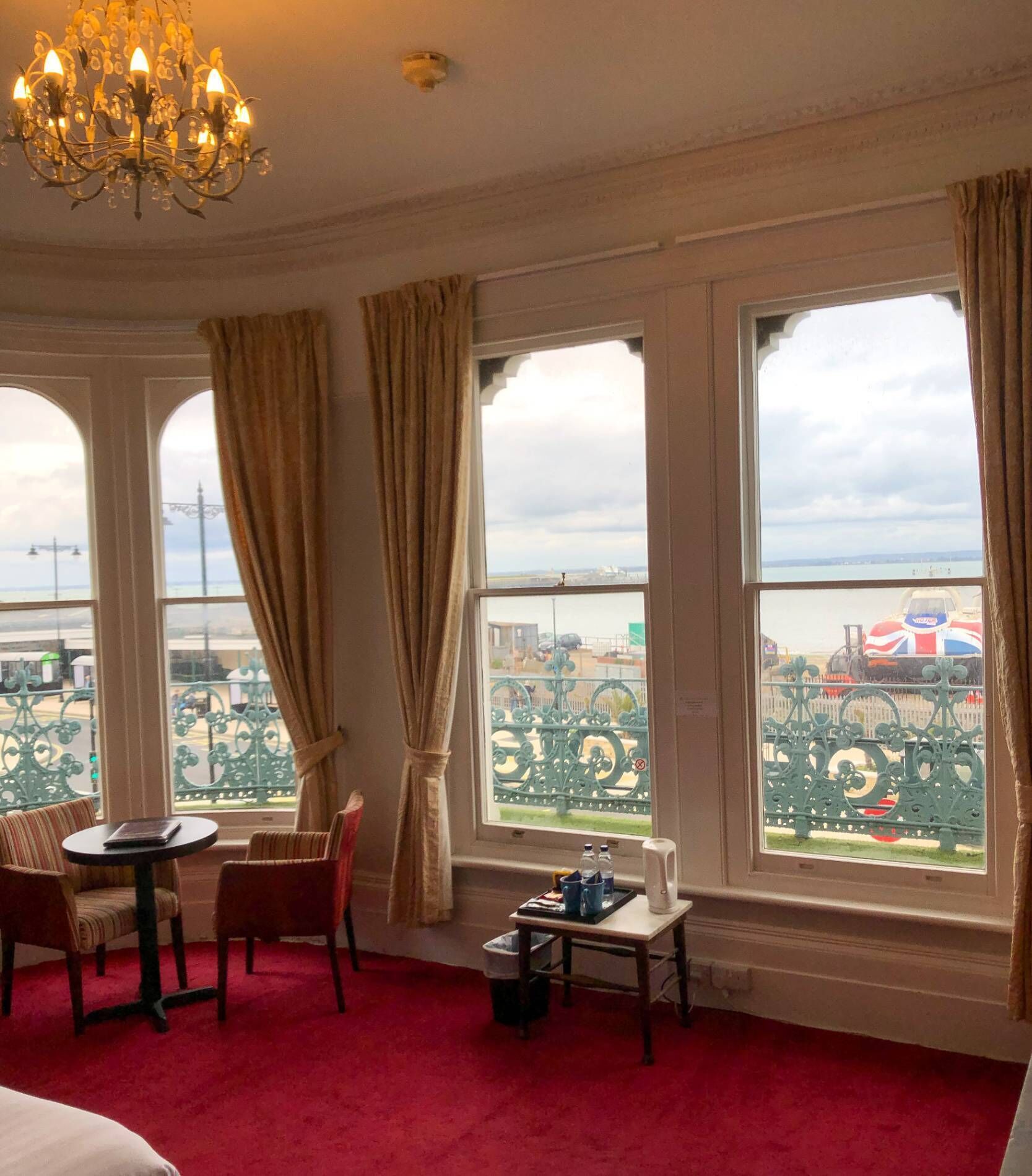 A room with a red carpet, a chandelier, a table with chairs, and large windows with curtains.