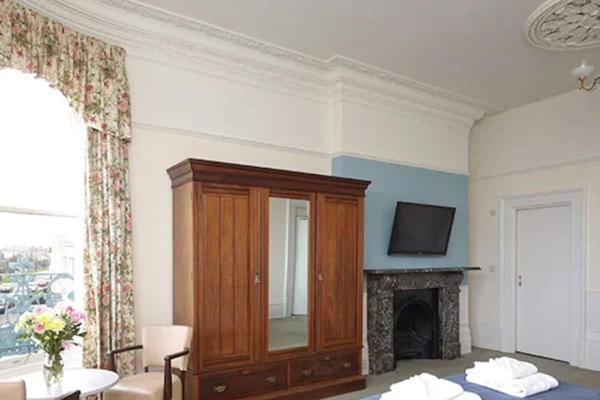 Quarto de hotel com cama, guarda-roupa de madeira, televisão, lareira, mesa de jantar com cadeiras e janela com cortinas.