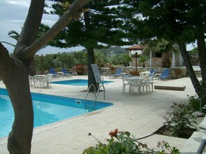 A pool area with lounge chairs and tables, surrounded by trees and potted plants.