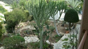 A garden with various plants, including a prominent cactus, stone pathways, and a view of a distant building. - Rantzo Holiday Apartments (Pissouri)