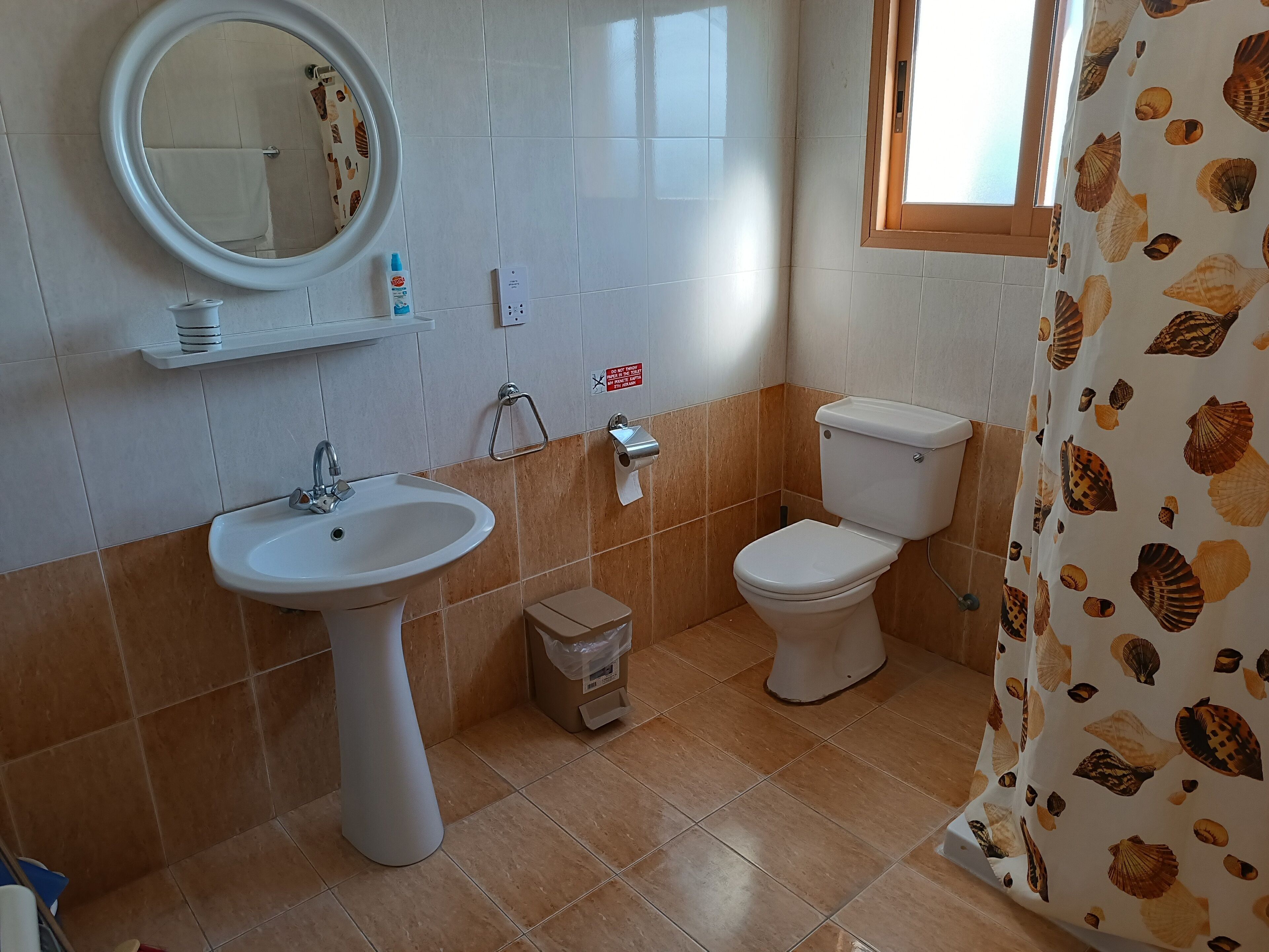 Ein Badezimmer mit Waschbecken, Toilette und Spiegel. Eine kleine Mülltonne und ein Ablagefach mit Pflegeprodukten sind ebenfalls vorhanden.