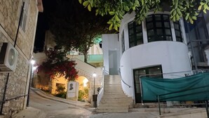 A nighttime view of a narrow street with cobblestone pavement, a white building with a curved facade, and a staircase leading to an upper level. - Kapsalia Holiday Villas (Pissouri)