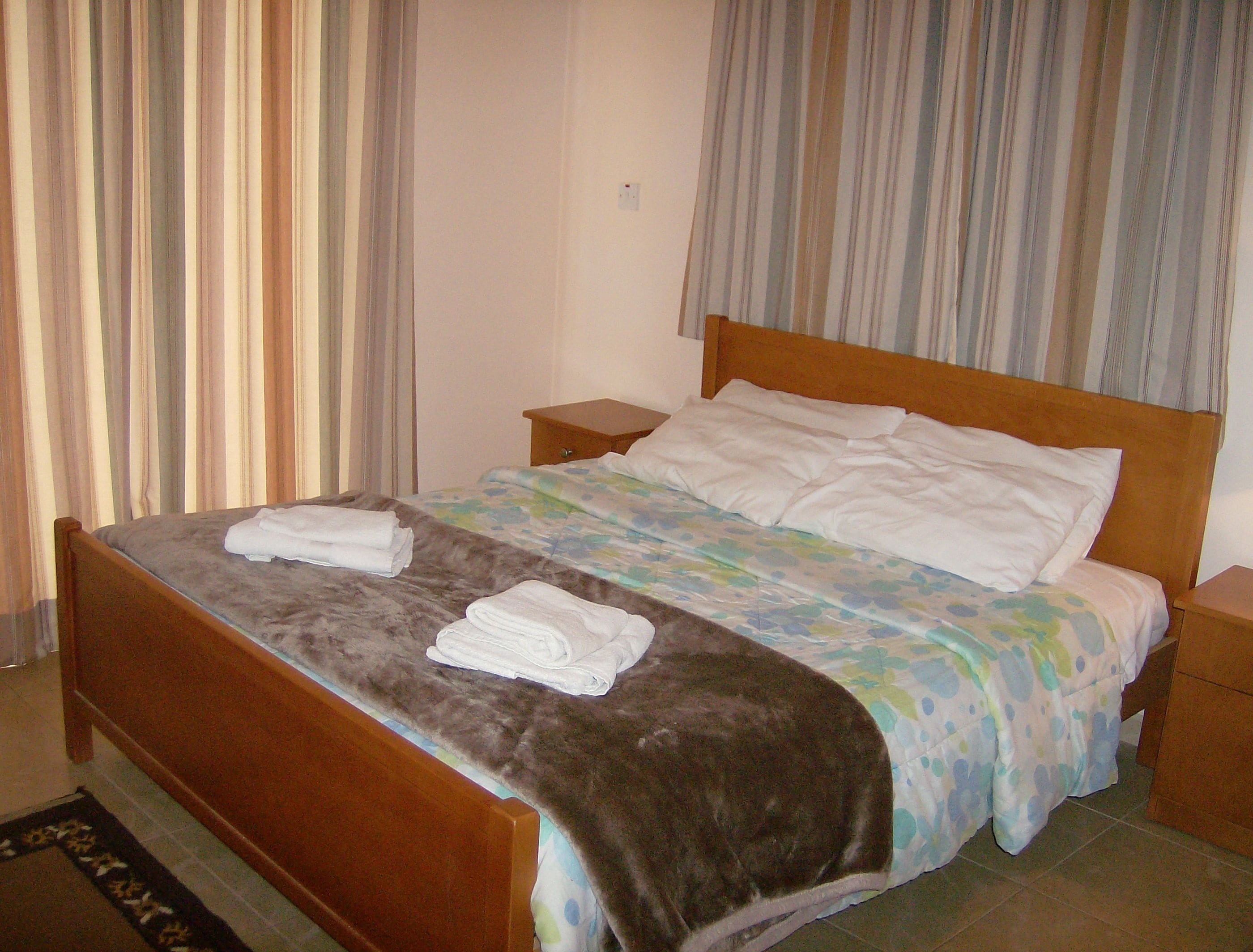 A neatly made bed with a brown headboard and a floral bedspread. There are two white pillows and a folded white towel on the bed. The room has a wooden nightstand beside the bed and a striped curtain in the background.