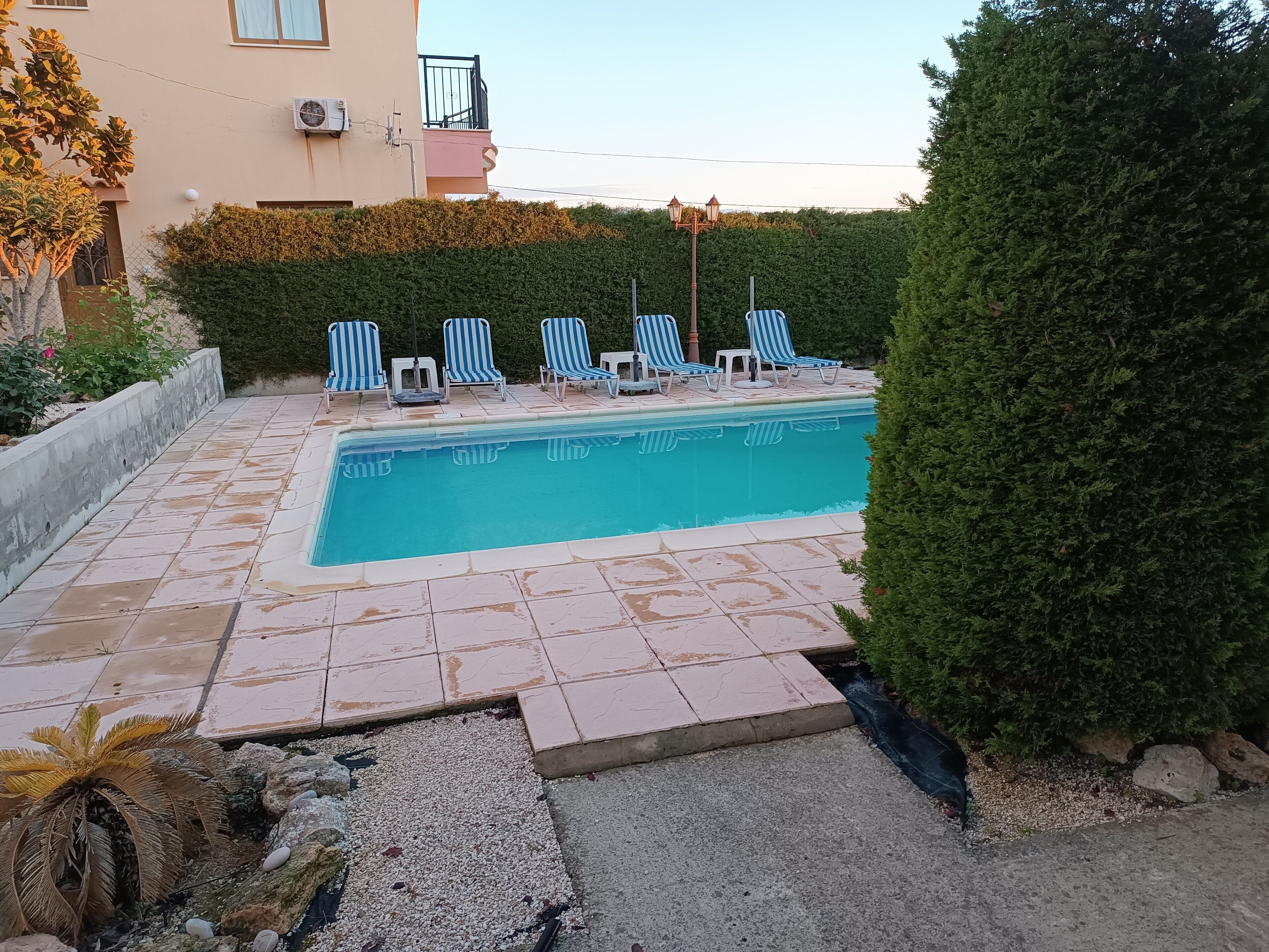Una piscina con sillas de lounge a rayas azules y blancas, rodeada por un patio de piedra y un seto.