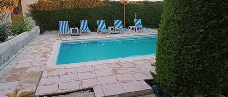 Una piscina con sillas de lounge a rayas azules y blancas, rodeada por un patio de piedra y un seto.
