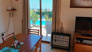 A room with a dining table, chairs, a TV, a heater, and a view of a swimming pool and greenery outside. - Kapsalia Holiday Villas (Pissouri)
