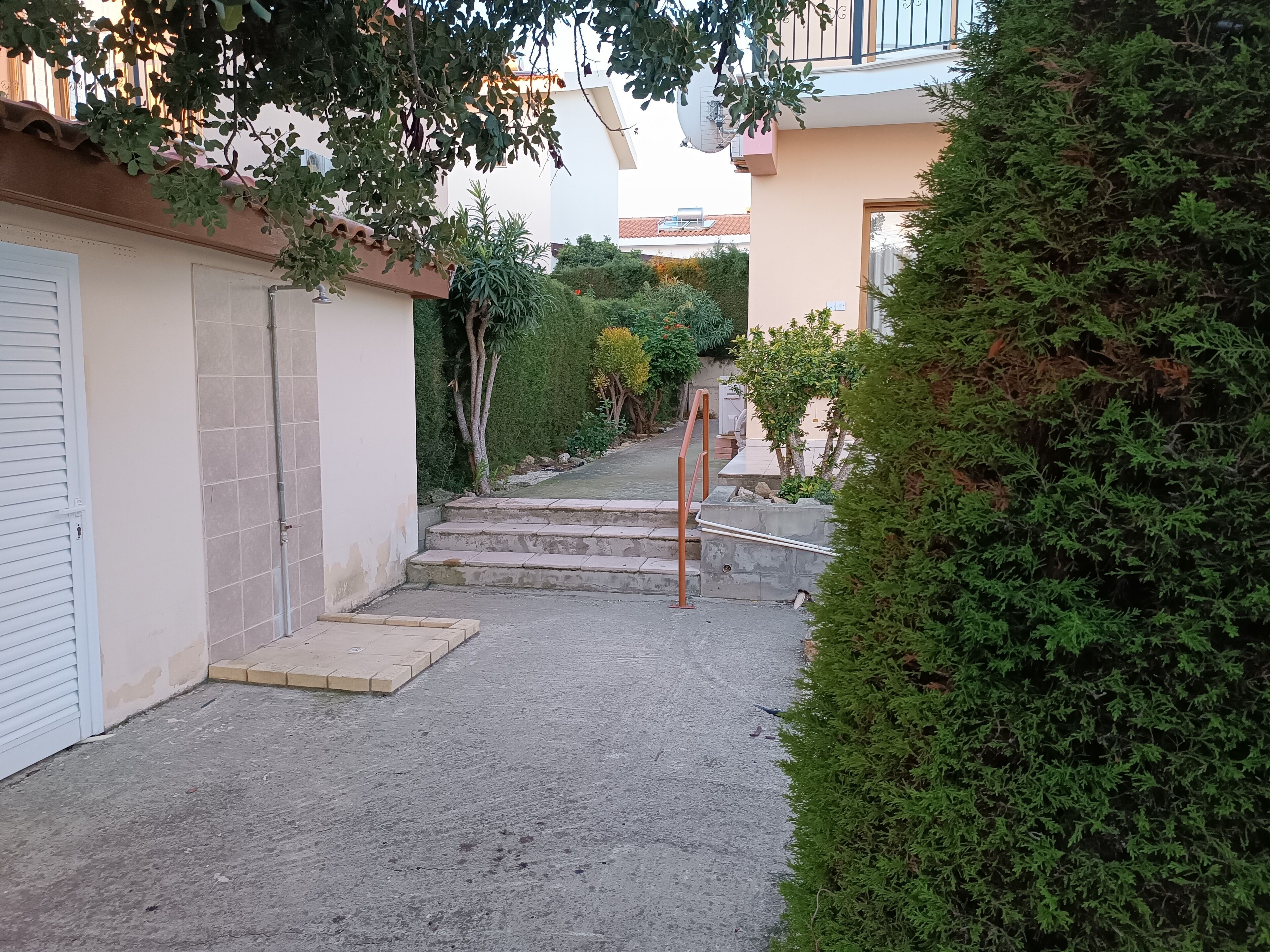 Un étroit sentier menant à l’entrée d’un bâtiment, avec un petit escalier et une rambarde en métal.