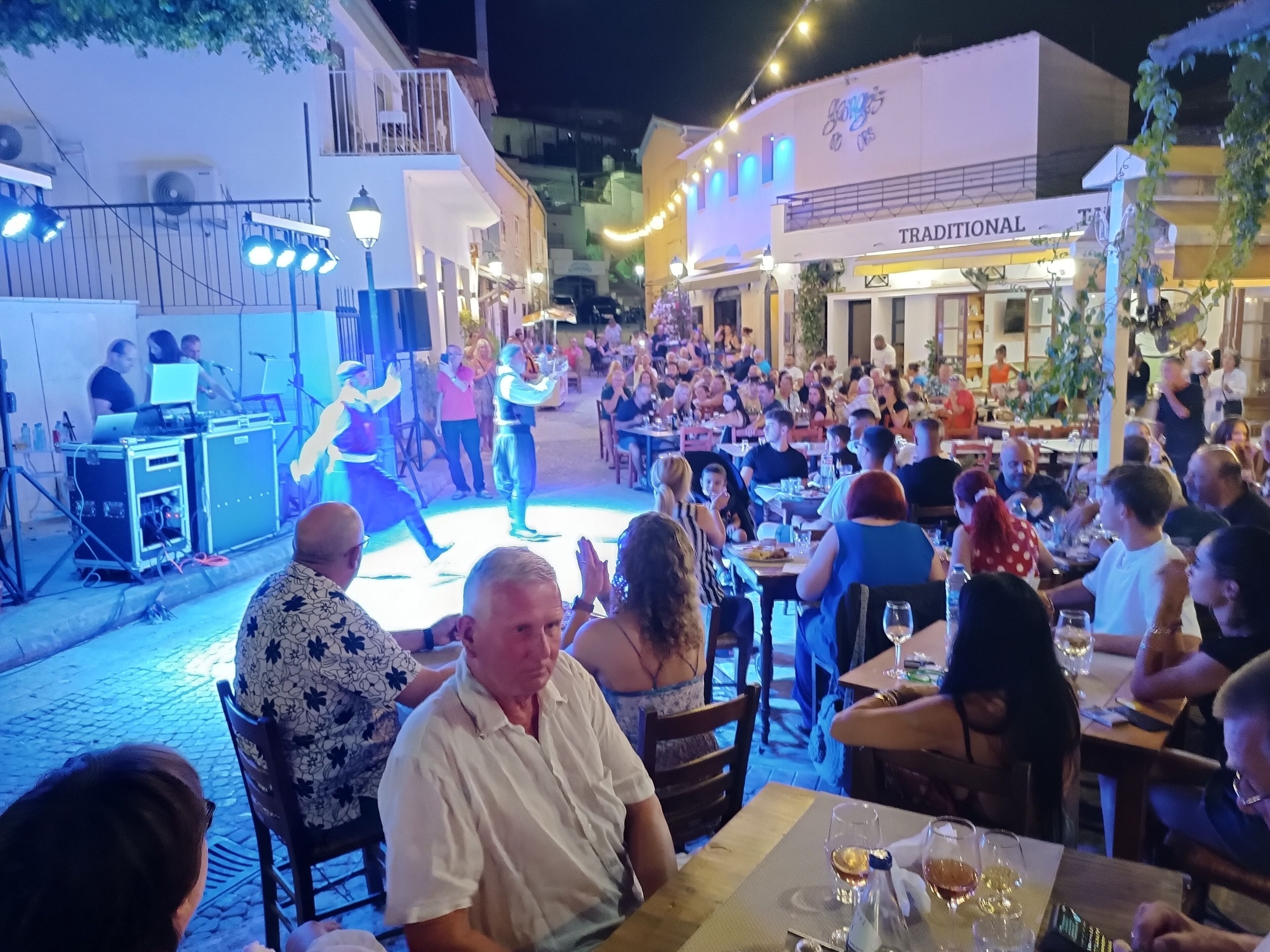 An outdoor dining area with tables and chairs, a stage with performers, and a building with a sign reading "TRADITIONAL".
