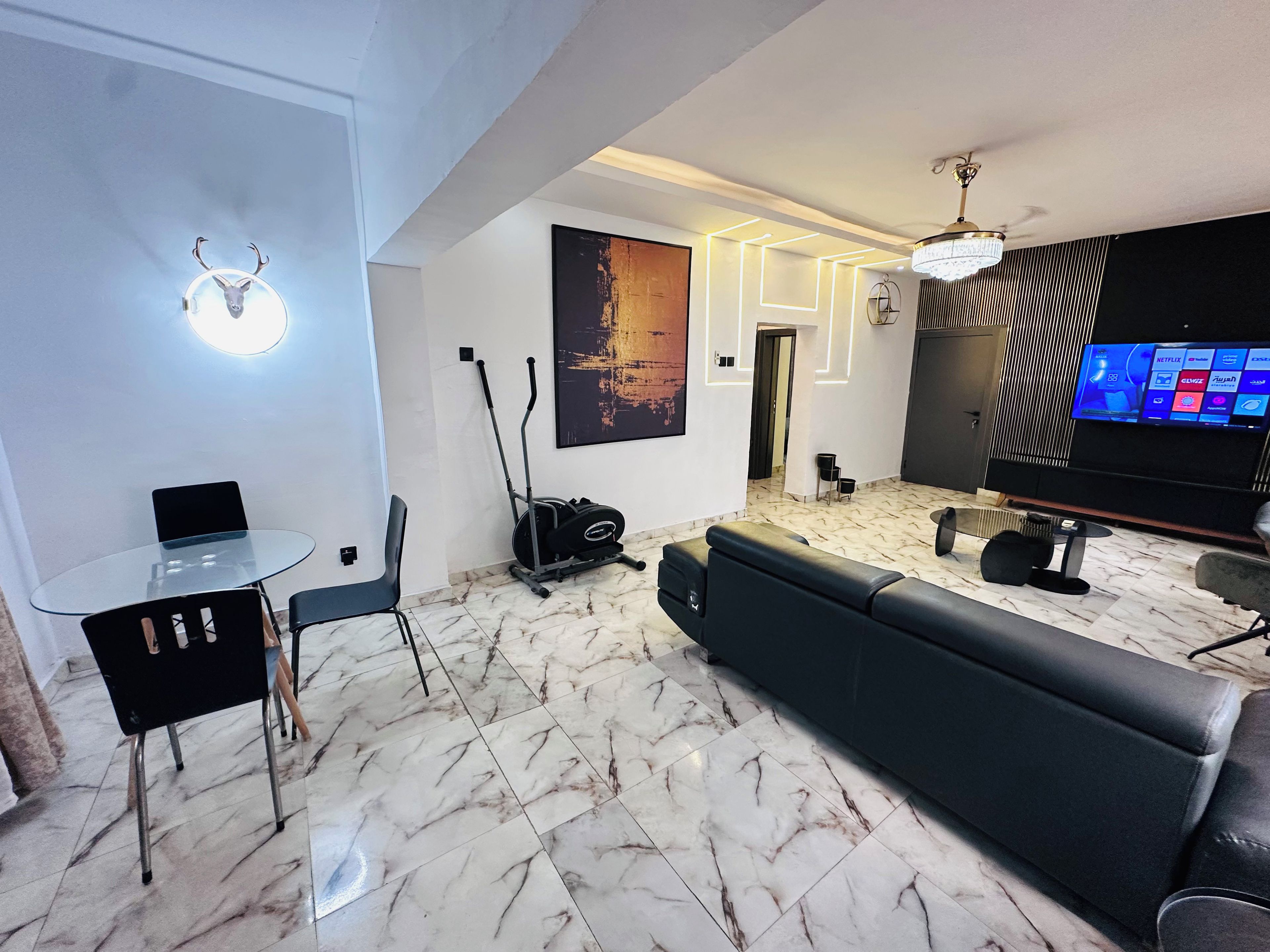 Un soggiorno moderno con tavolo in vetro, sedie nere, un divano nero, un tapis roulant e una TV che mostra diversi servizi di streaming.