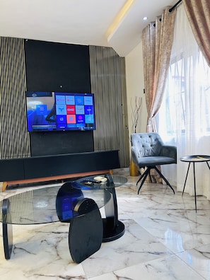 A modern living room with a flat-screen TV mounted on a black wall, a glass coffee table with a remote control, a grey sofa, and a small side table. - C.G. Apartments (Lagos)